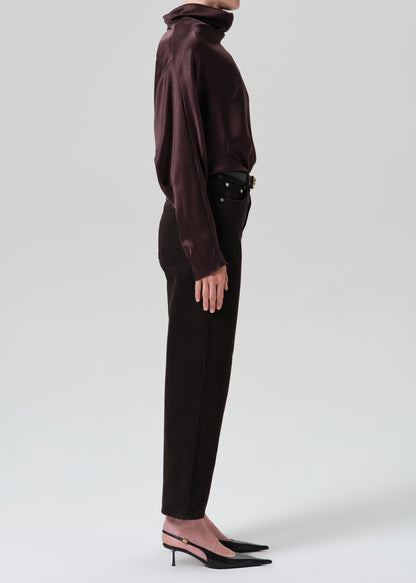 A model shown in profile wearing a dark plum satin long-sleeve turtleneck top, dark brown trousers, a black belt, and black pointed-toe slingback heels against a grey background.