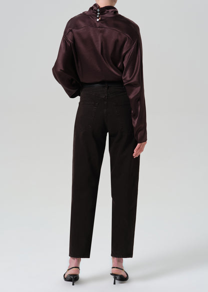 A model shown from the back wearing a dark plum satin long-sleeve blouse with a high mock neck and decorative buttons, paired with dark brown trousers and black heels, against a grey background.