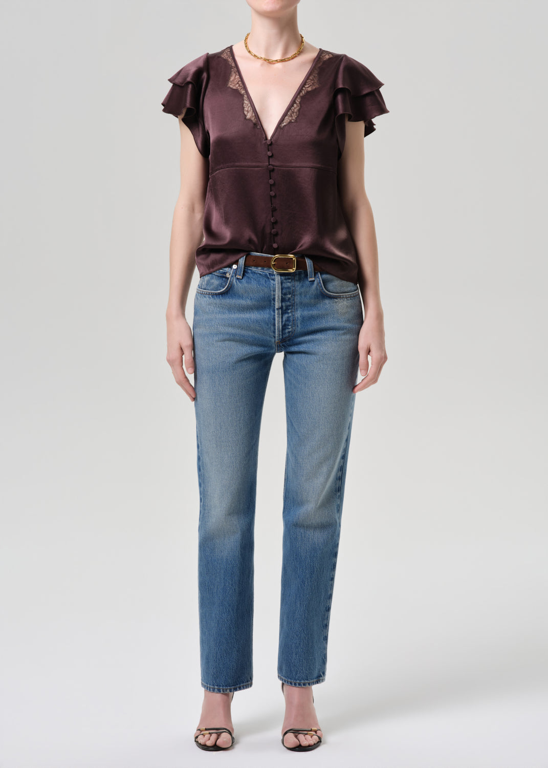 Woman wearing a brown satin blouse with ruffled sleeves and blue jeans on a grey background