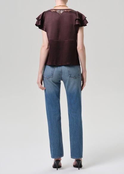 Person wearing a brown top with ruffled sleeves and blue jeans on a grey background
