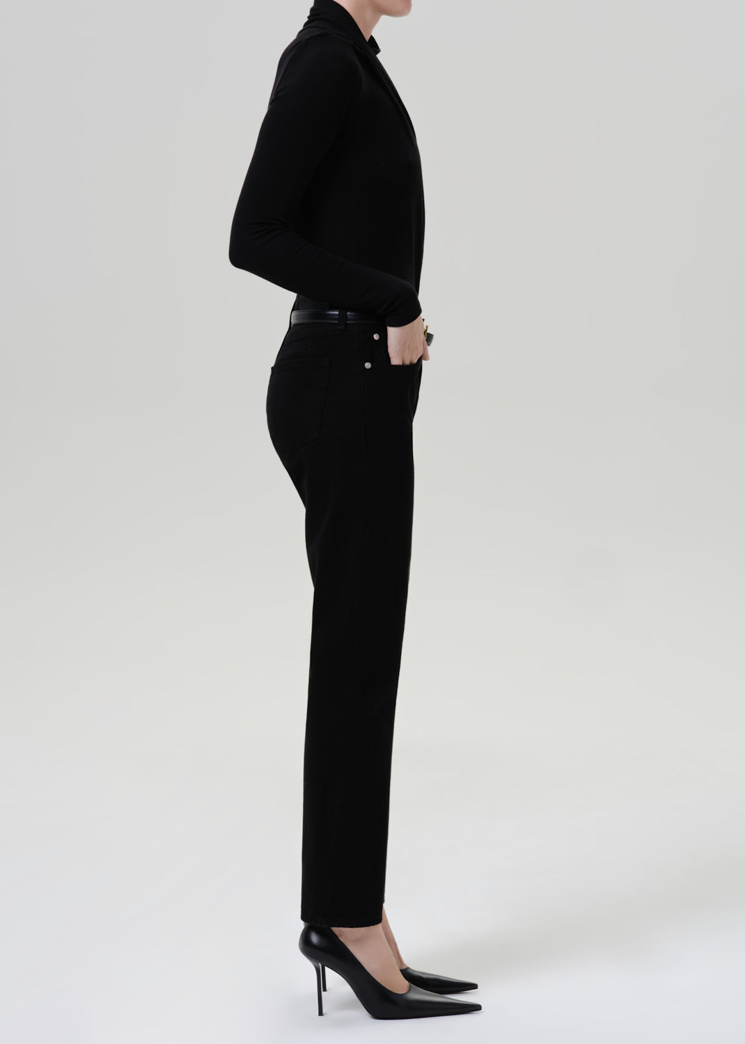 Person wearing a black fitted turtleneck with black straight leg pants viewed from the side on a grey background
