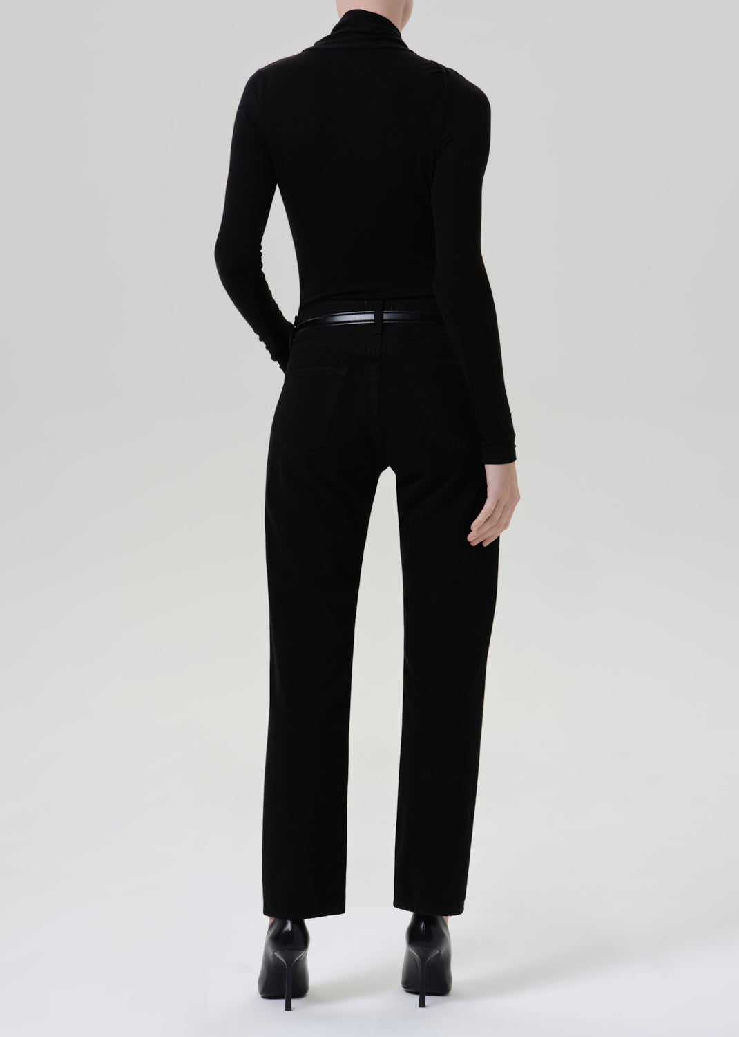 Person wearing a black fitted turtleneck with black straight leg pants viewed from the back on a grey background