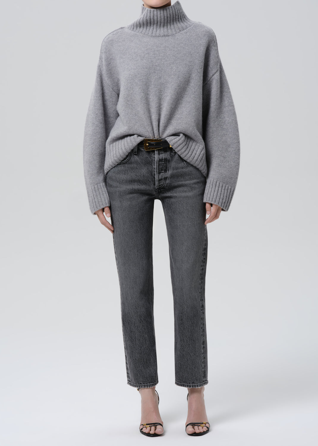 Person wearing a grey turtleneck sweater with grey straight leg jeans on a grey background