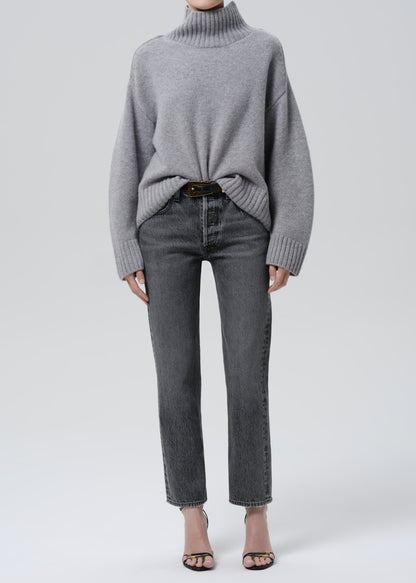 Person wearing a grey turtleneck sweater with grey straight leg jeans on a grey background