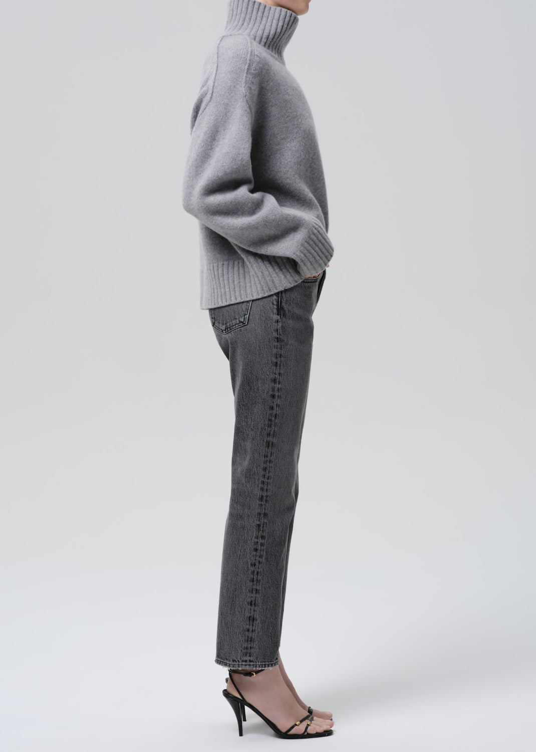 A side-profile view of a model wearing a light gray ribbed turtleneck sweater, dark gray slim-fit jeans with subtle vertical whiskering, and black strappy high-heeled sandals against a grey background.