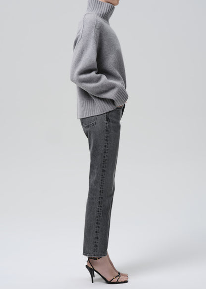 A side-profile view of a model wearing a light gray ribbed turtleneck sweater, dark gray slim-fit jeans with subtle vertical whiskering, and black strappy high-heeled sandals against a grey background.