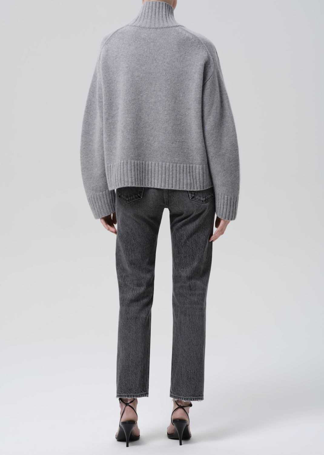 Person wearing a grey turtleneck sweater with grey straight leg jeans viewed from the back on a grey background