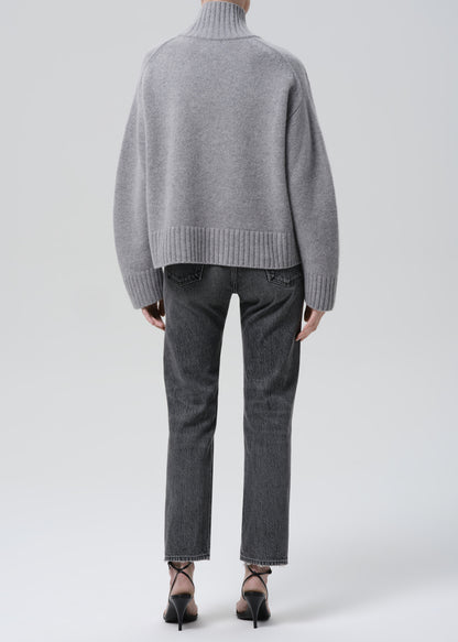 Person wearing a grey turtleneck sweater with grey straight leg jeans viewed from the back on a grey background
