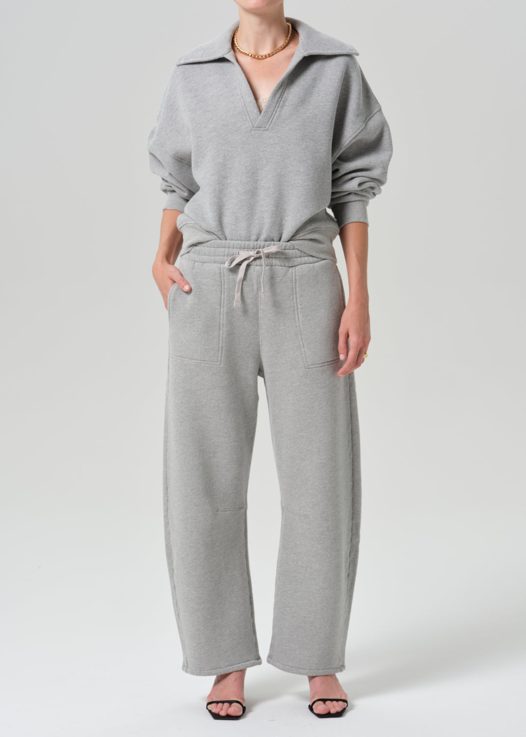 A model wearing a light grey two-piece loungewear set consisting of a half-zip sweatshirt and matching wide-leg sweatpants, photographed against a grey background.