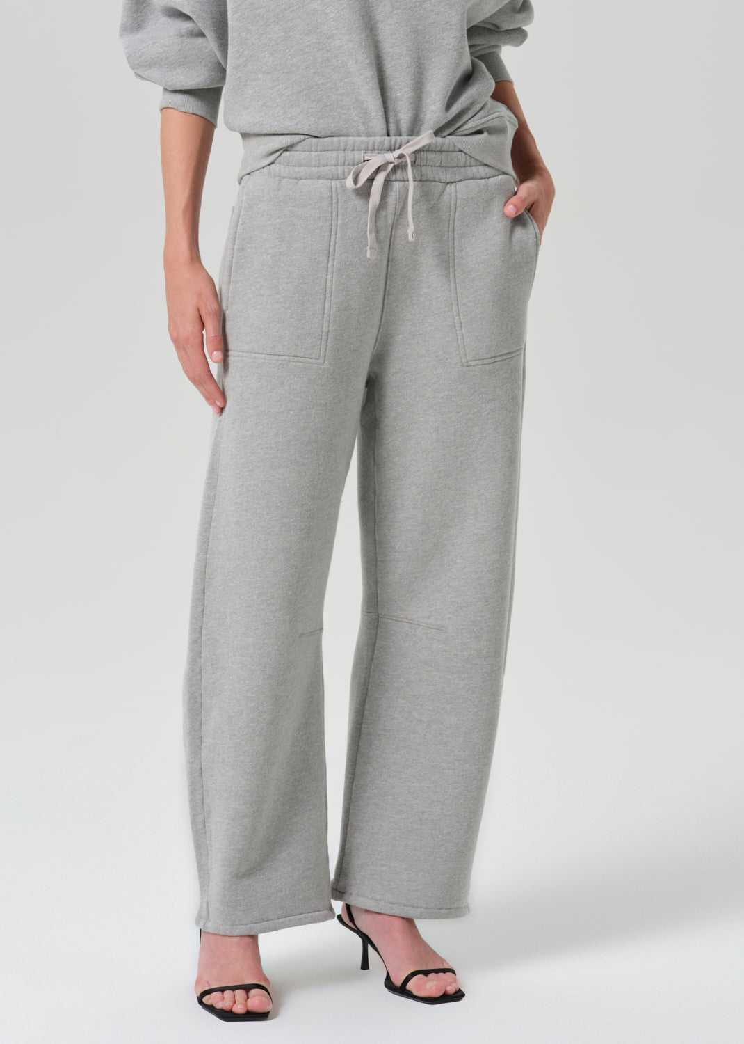A person wearing light heather grey wide-leg sweatpants with a drawstring waist, paired with a matching gray sweatshirt and black heeled sandals, photographed against a grey background.