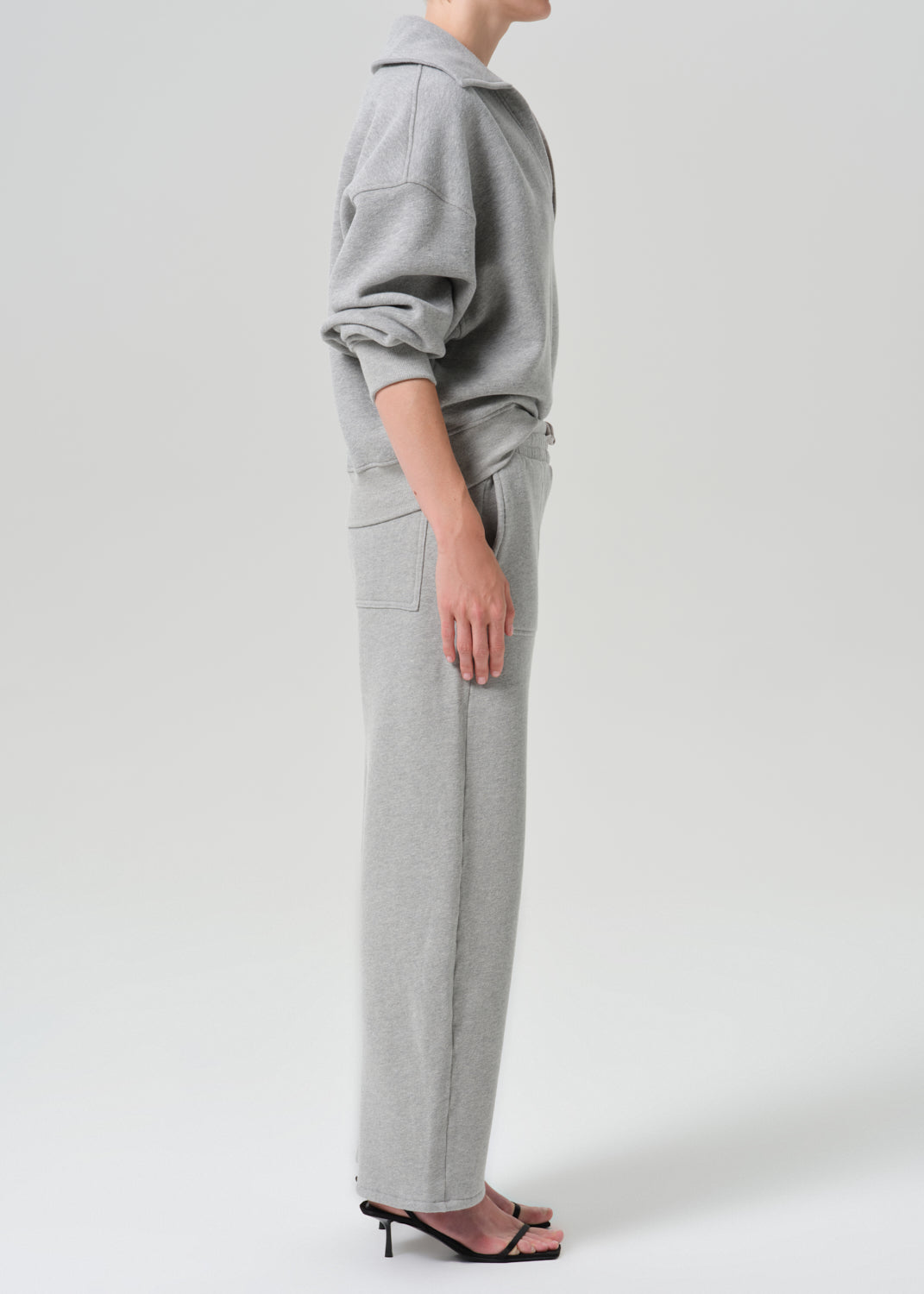 A model shown from the side wearing a light grey heathered sweatshirt and matching wide-leg sweatpants, paired with black heeled sandals, against a grey background.