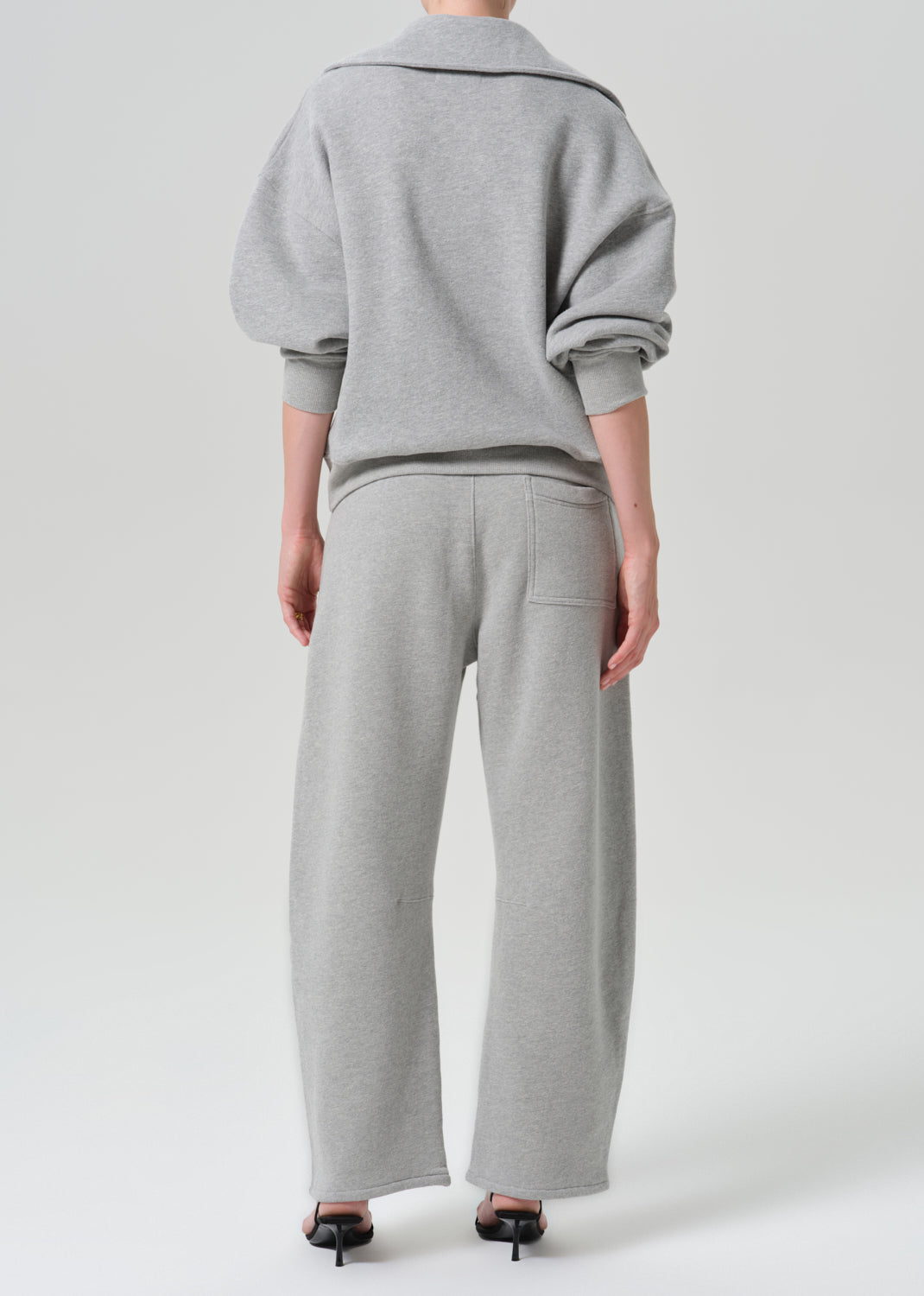 A model viewed from the back wearing a light grey heathered sweatshirt with voluminous sleeves and matching wide-leg sweatpants, paired with black high-heeled sandals against a grey background.