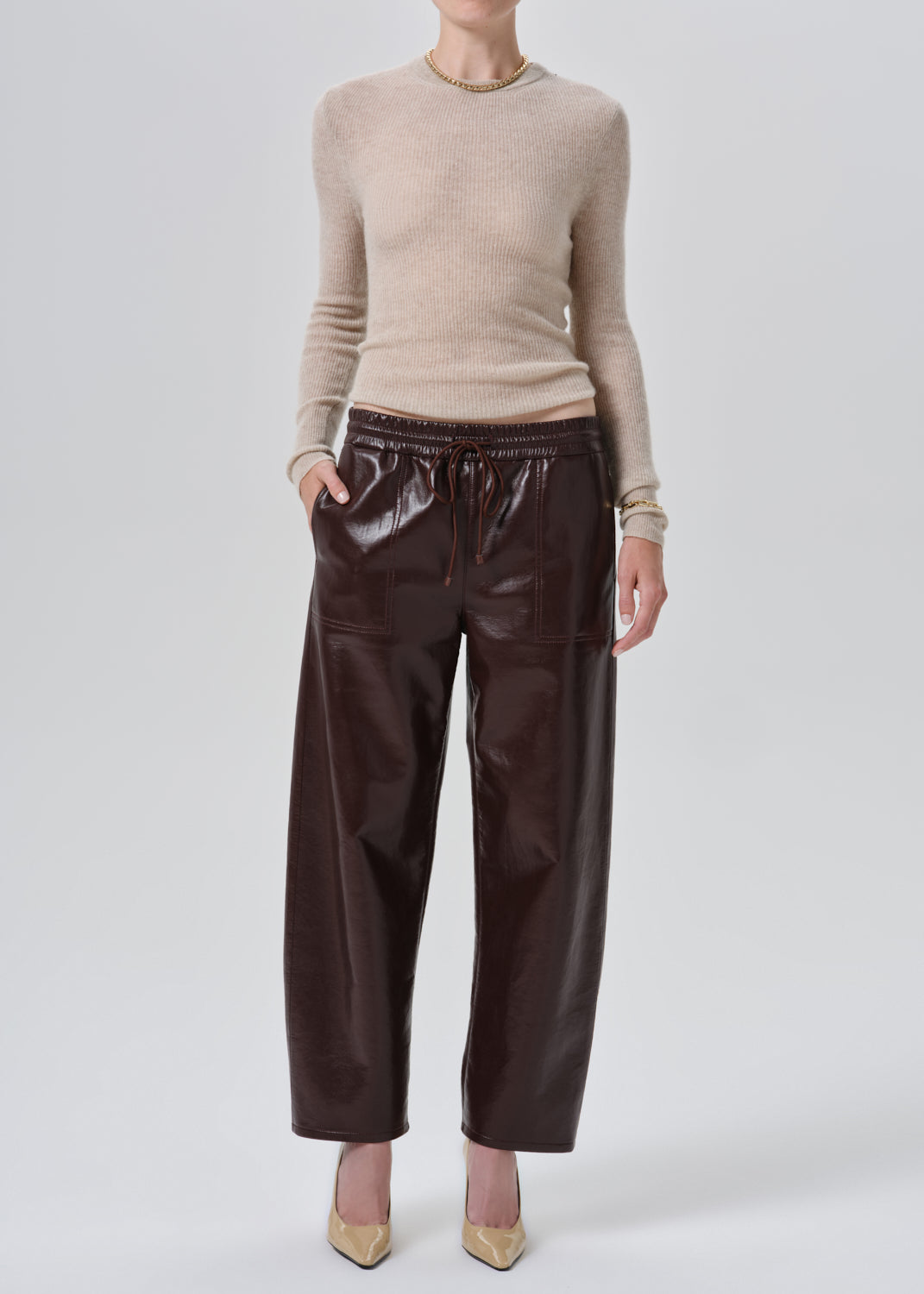 A model wearing a beige knit mock-neck sweater and dark brown wide-leg faux leather pants, standing against a grey studio background.
