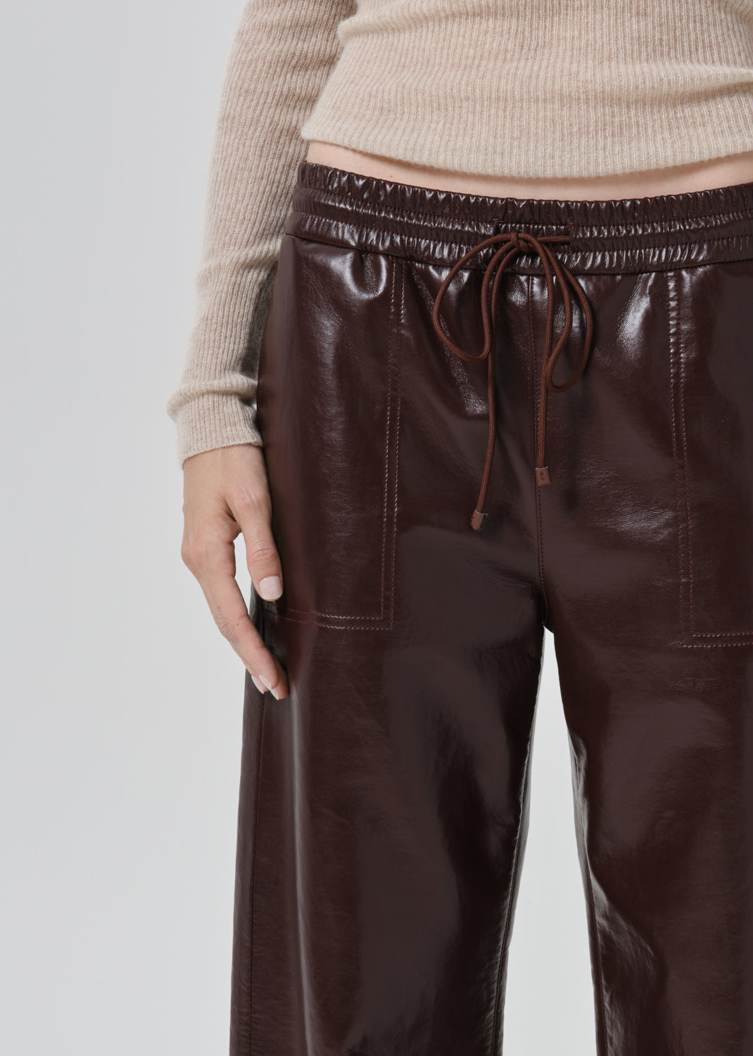 Close-up of a person wearing dark brown faux-leather wide-leg pants with an elastic drawstring waistband, paired with a beige ribbed sweater, against a grey background.