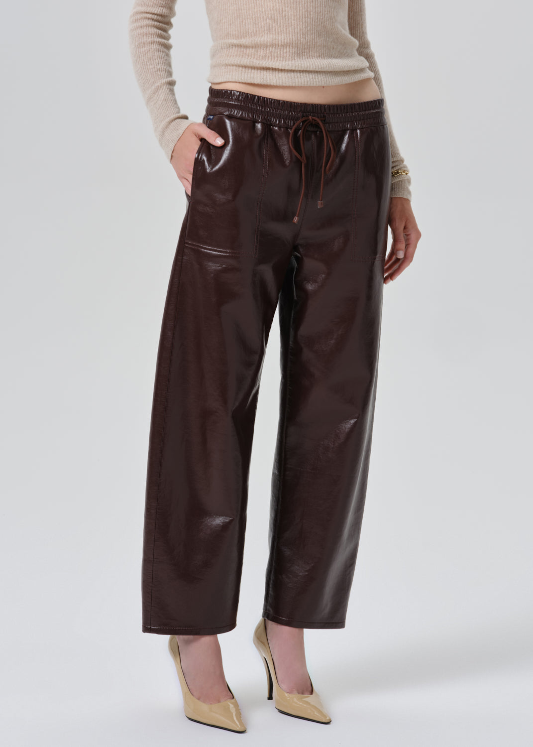 A model wearing dark brown wide-leg faux leather pants with an elastic drawstring waist, paired with a beige ribbed sweater and beige pointed-toe heels, against a grey background.