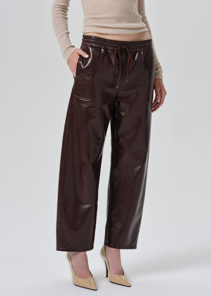 A model wearing dark brown wide-leg faux leather pants with an elastic drawstring waist, paired with a beige ribbed sweater and beige pointed-toe heels, against a grey background.