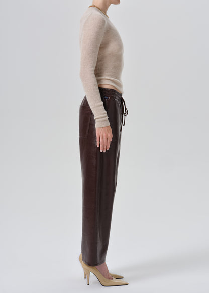 A model shown in profile wearing dark brown leather trousers, a beige ribbed knit long-sleeve crop top, and beige pointed-toe heels against a grey background.