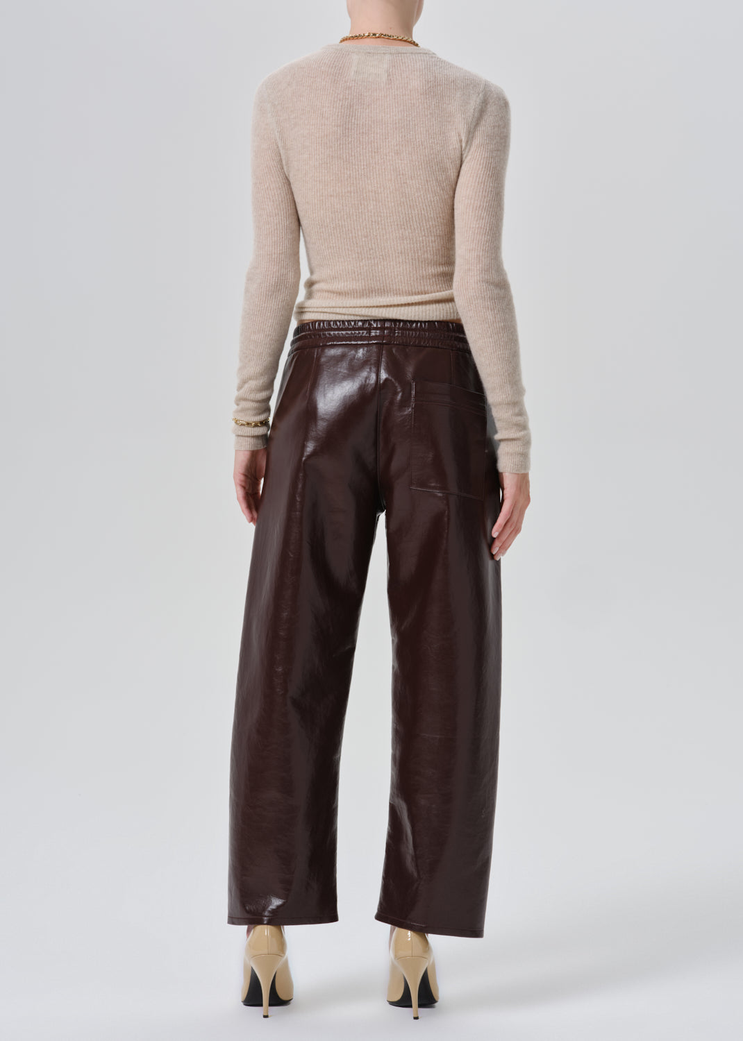 A model shown from the back wearing dark brown wide-leg faux leather trousers, a beige knit sweater, and beige high heels against a grey background.