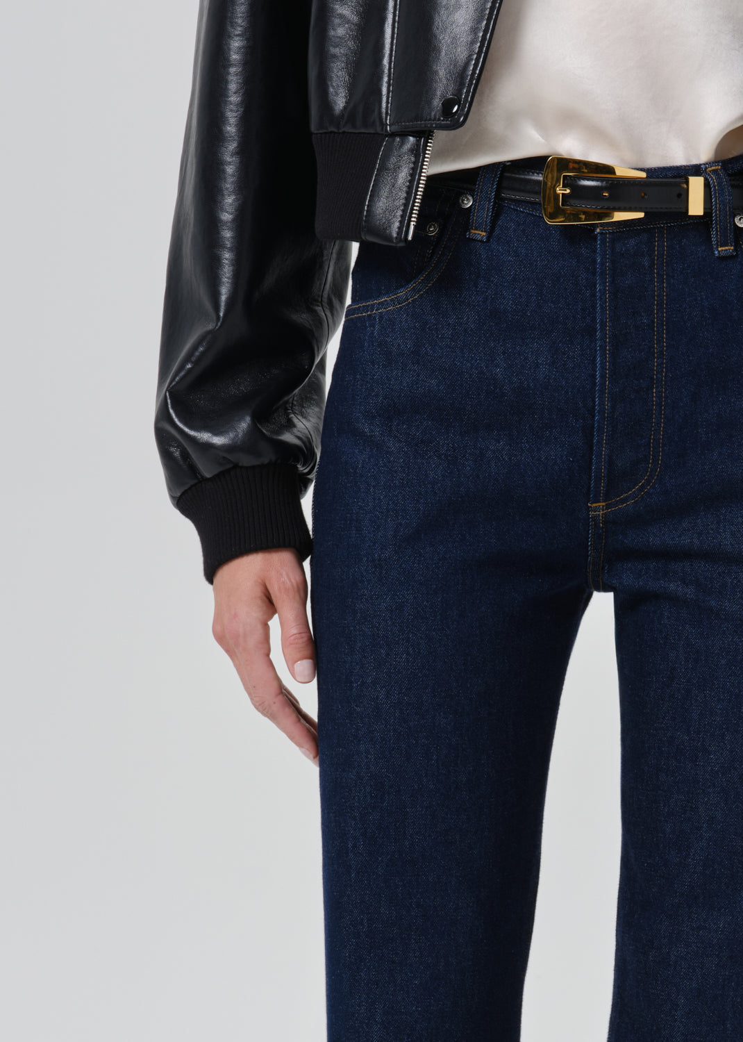 Close-up of a person wearing dark blue denim jeans with a black belt featuring a gold buckle, styled with a black faux-leather jacket and a cream top against a grey background.