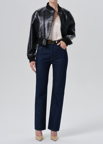 A model wearing a black leather bomber jacket, dark blue straight-leg jeans, a cream top, and beige heels, standing against a grey background.
