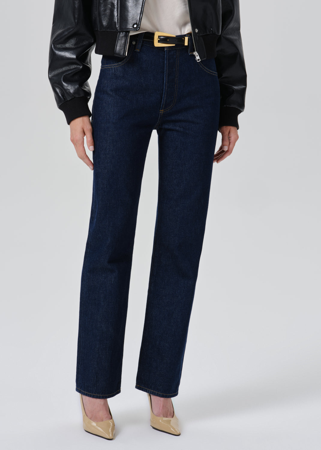A model wearing dark blue straight-leg denim jeans with a gold-tone belt, styled with a black leather jacket, beige top, and beige pointed-toe heels against a grey background.