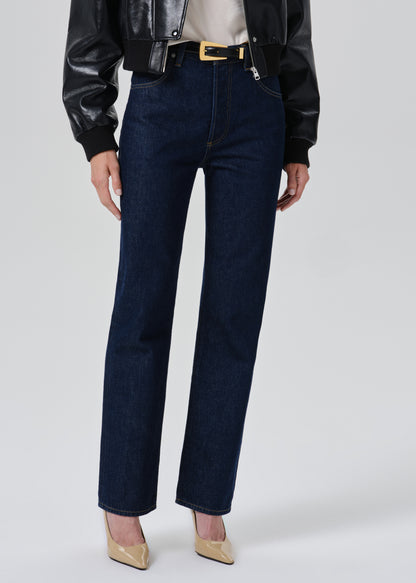A model wearing dark blue straight-leg denim jeans with a gold-tone belt, styled with a black leather jacket, beige top, and beige pointed-toe heels against a grey background.