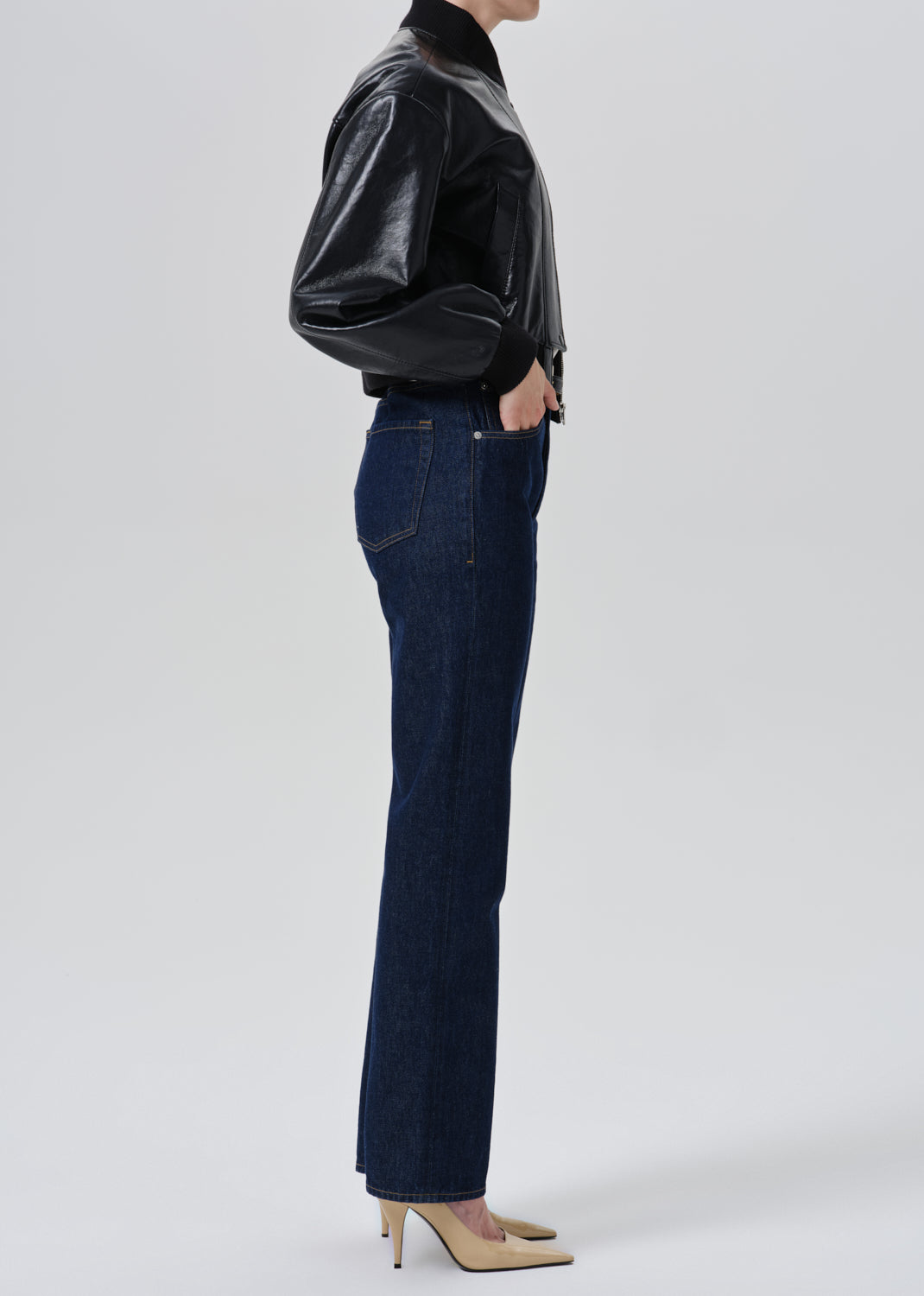 A side-profile studio shot of a model wearing dark blue straight-leg jeans, a black faux-leather jacket, and beige heels against a grey background.