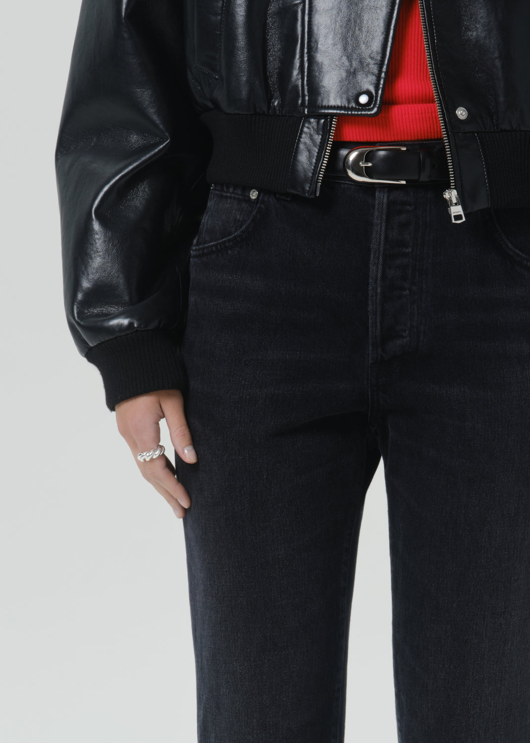 Close-up of a person wearing a black faux-leather moto jacket, black jeans, a black belt, and a red top, with a silver ring on one hand, against a grey background.