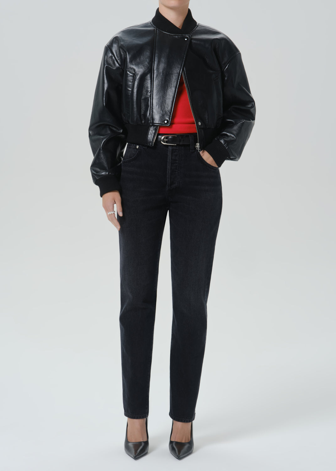 A model wearing a black leather bomber jacket, black skinny jeans, a red top, and black pointed-toe heels, photographed against a grey background.