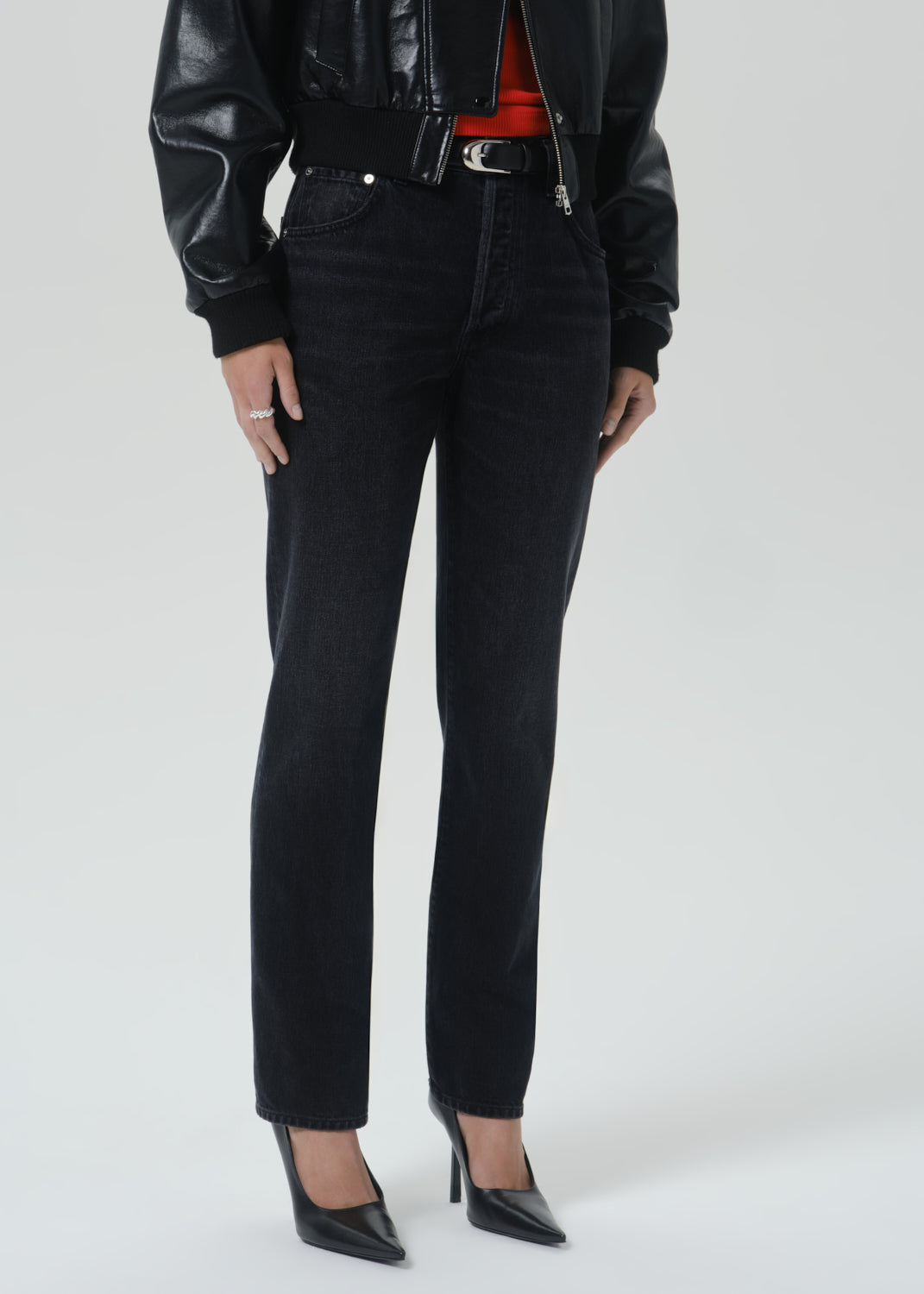 A model wearing black slim-fit jeans with a black leather jacket, red top, and black pointed-toe heels, shown from the chest down on a grey background.