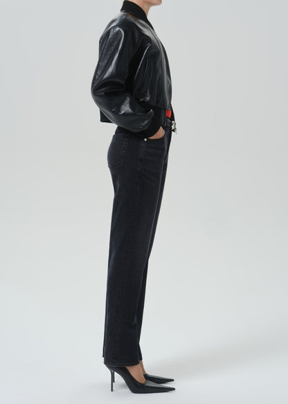 A model shown from the side wearing a black glossy cropped jacket, black jeans, and black pointed-toe heels against a grey background.