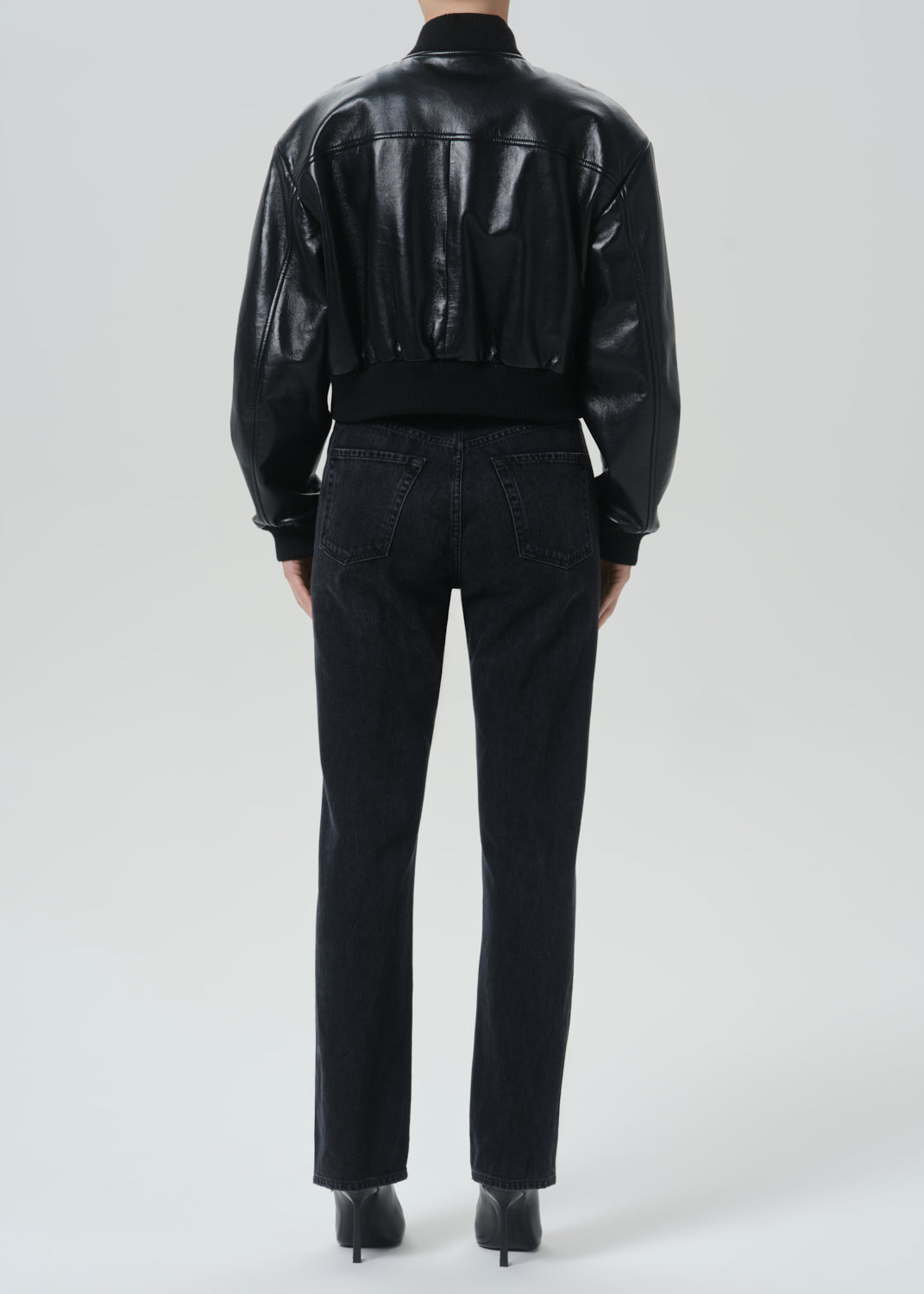 A model shown from the back wearing a black cropped faux-leather bomber jacket with a ribbed waistband, paired with black jeans and black heels, against a grey studio background.