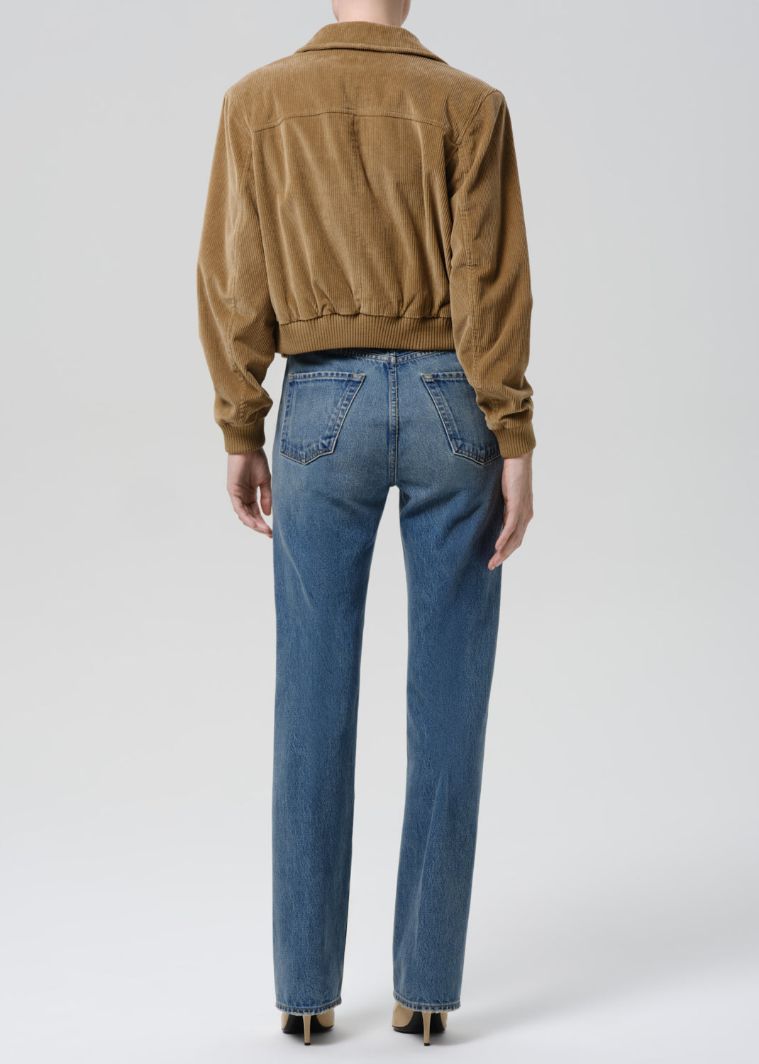 Person wearing a brown corduroy jacket and blue jeans on a grey background