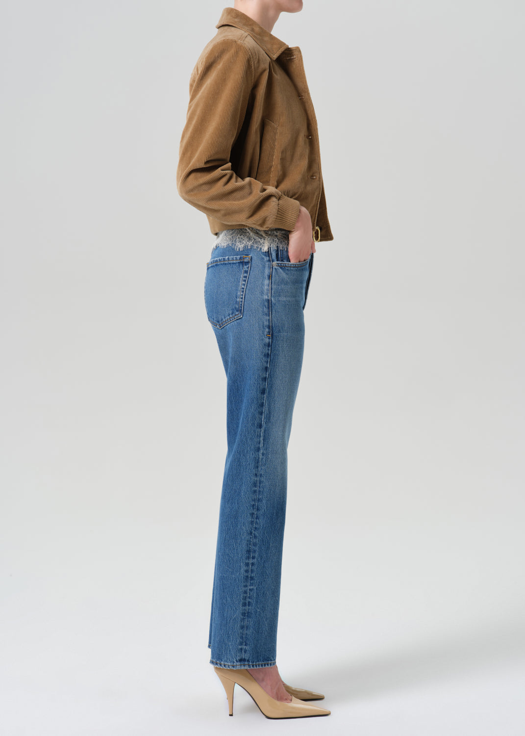 Person wearing brown jacket on a white lace top and blue pants on a grey background