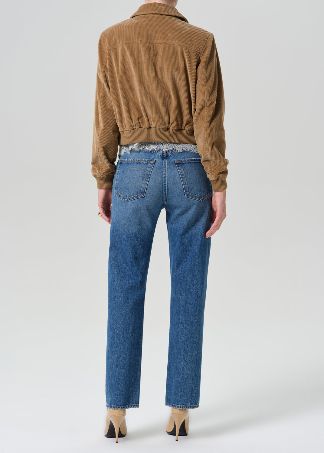 Person wearing brown jacket on a white lace top and blue pants on a grey background