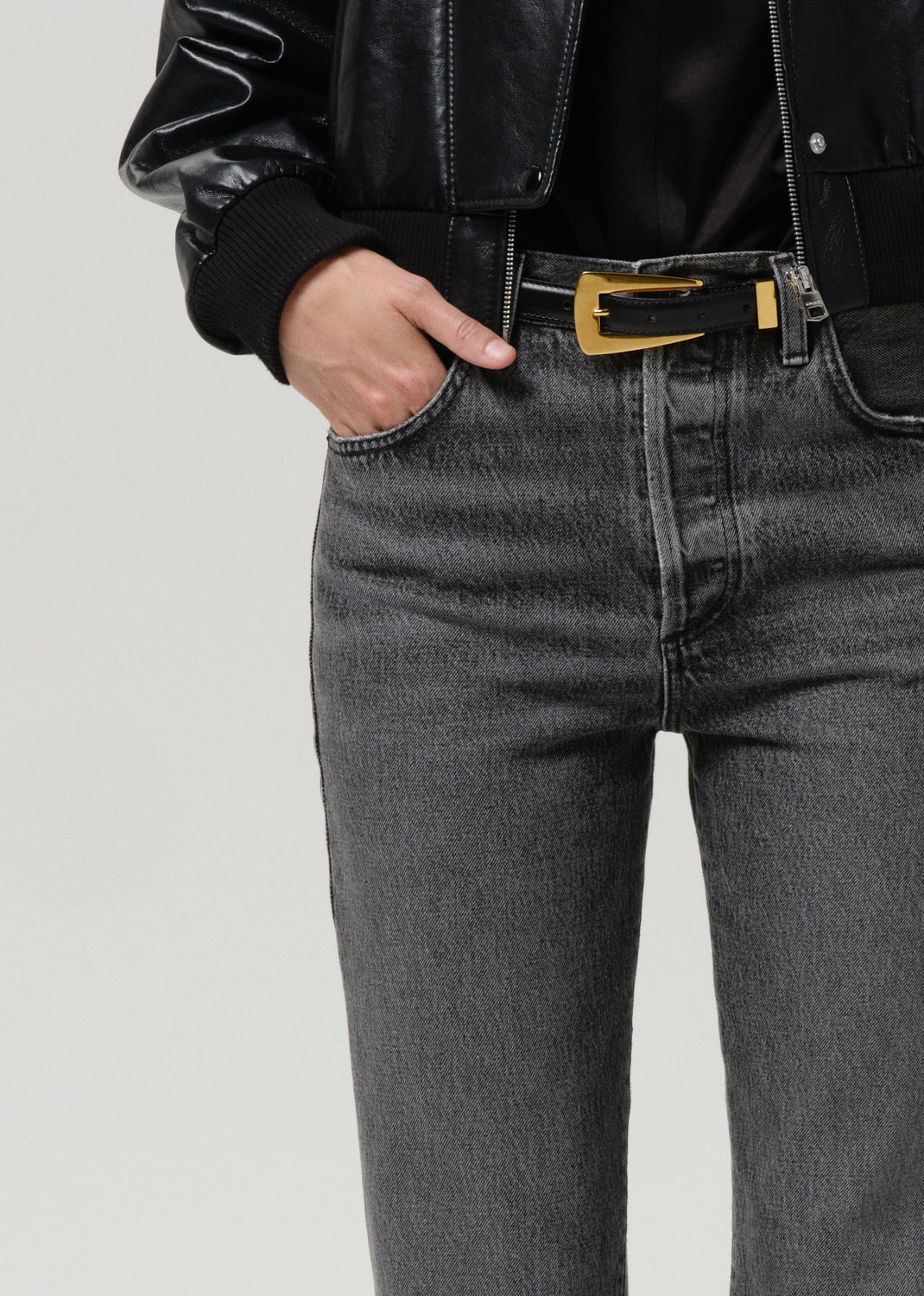 Person wearing a black leather jacket and gray jeans with a gold belt buckle on a grey background