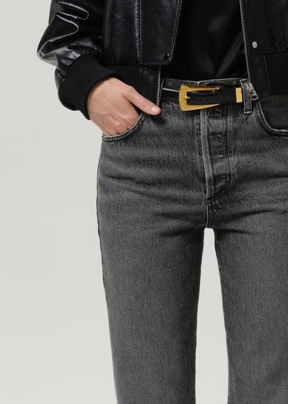 Person wearing a black leather jacket and grey jeans with a gold belt buckle on a grey background.