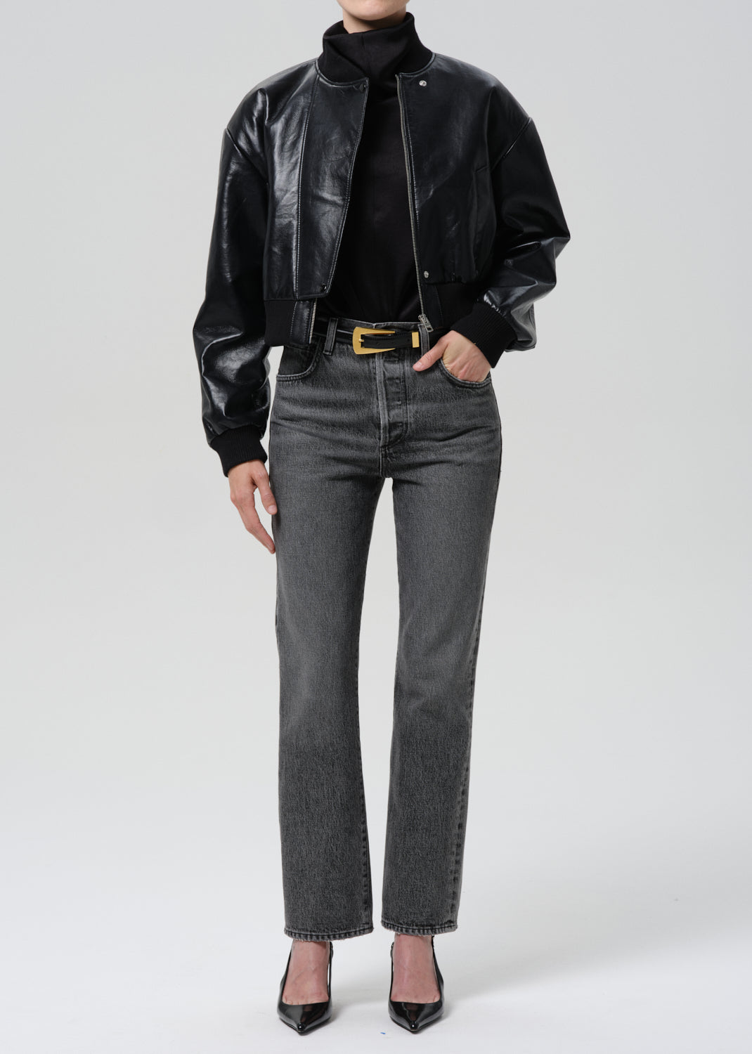 Person wearing a black leather jacket, black turtleneck, gray jeans, and black shoes on a grey background