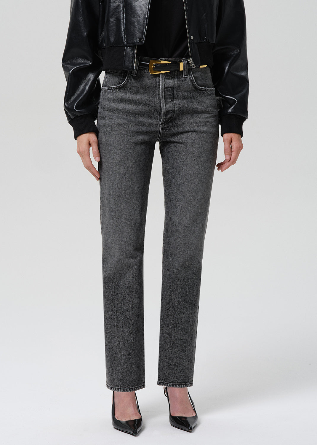 A model wearing a pair of dark grey straight-leg denim jeans, styled with a black leather jacket, black top, gold belt, and black pointed-toe heels against a grey background.