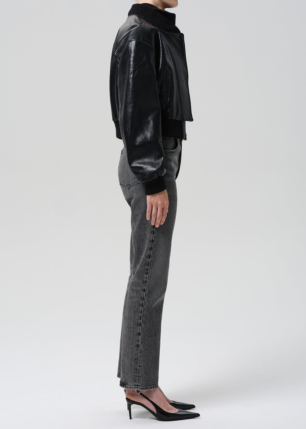 A model shown from the side wearing a black cropped faux-leather jacket, grey wide-leg jeans, and black pointed-toe heels against a grey background.