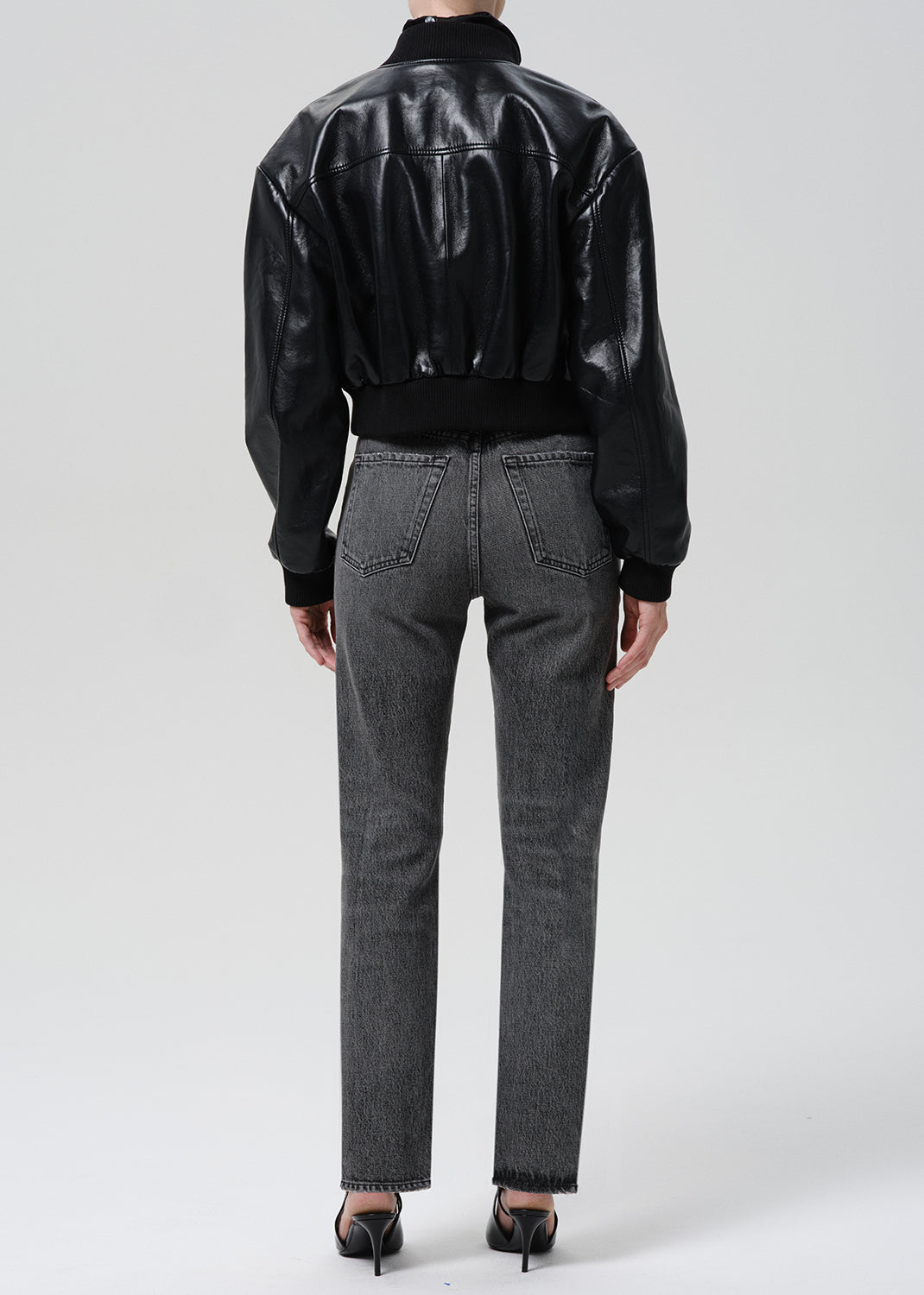 A model shown from the back wearing a black leather bomber jacket and dark grey straight-leg jeans, paired with black high heels, against a grey background.