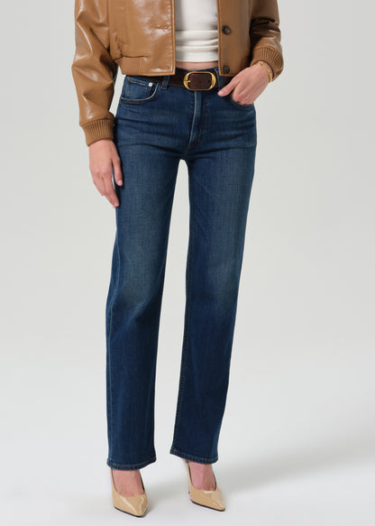 Person wearing a tan leather bomber jacket with dark indigo straight leg jeans on a grey background