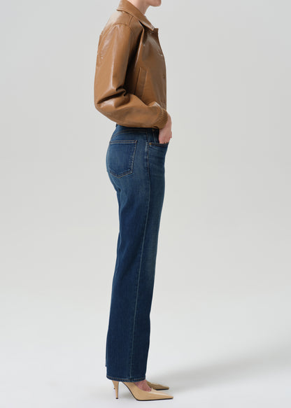 Person wearing a tan leather bomber jacket with dark indigo straight leg jeans viewed from the side on a grey background