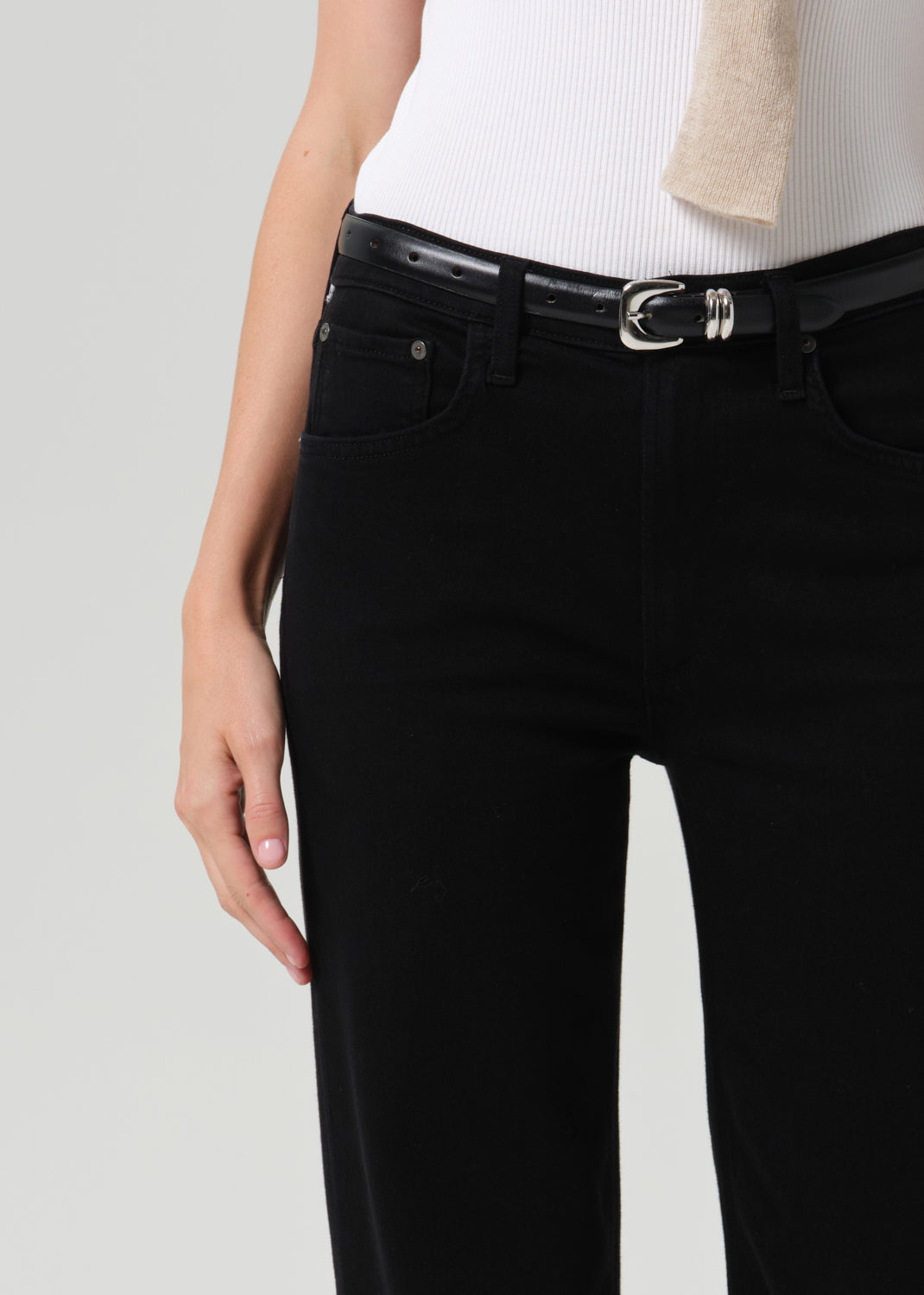 Close-up of black straight leg jeans with a white sleeveless knit top and sweater draped over the shoulders on a grey background