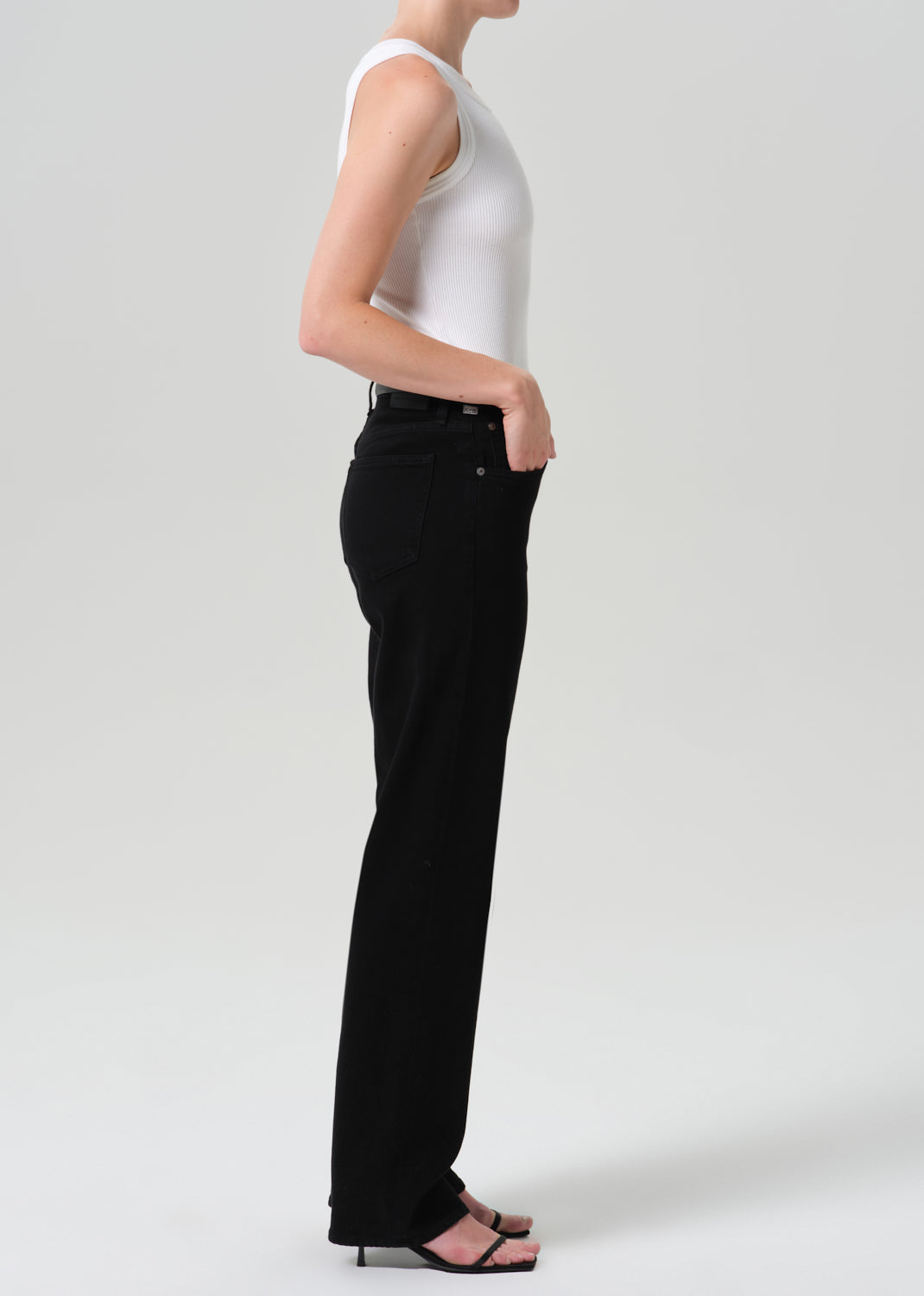 Person wearing a white sleeveless knit top with a sweater draped over the shoulders and black straight leg jeans viewed from the side on a grey background