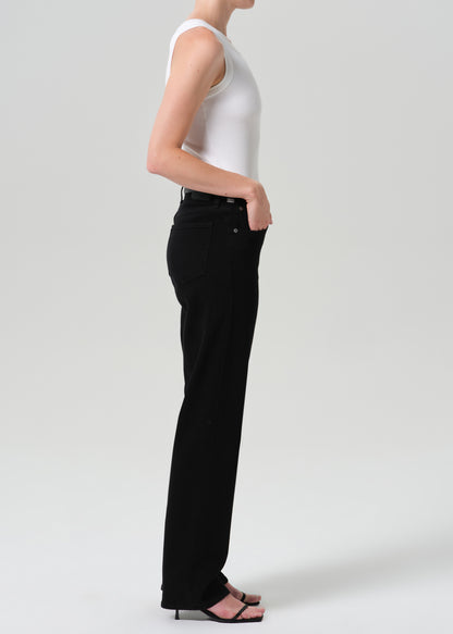 Person wearing a white sleeveless knit top with a sweater draped over the shoulders and black straight leg jeans viewed from the side on a grey background