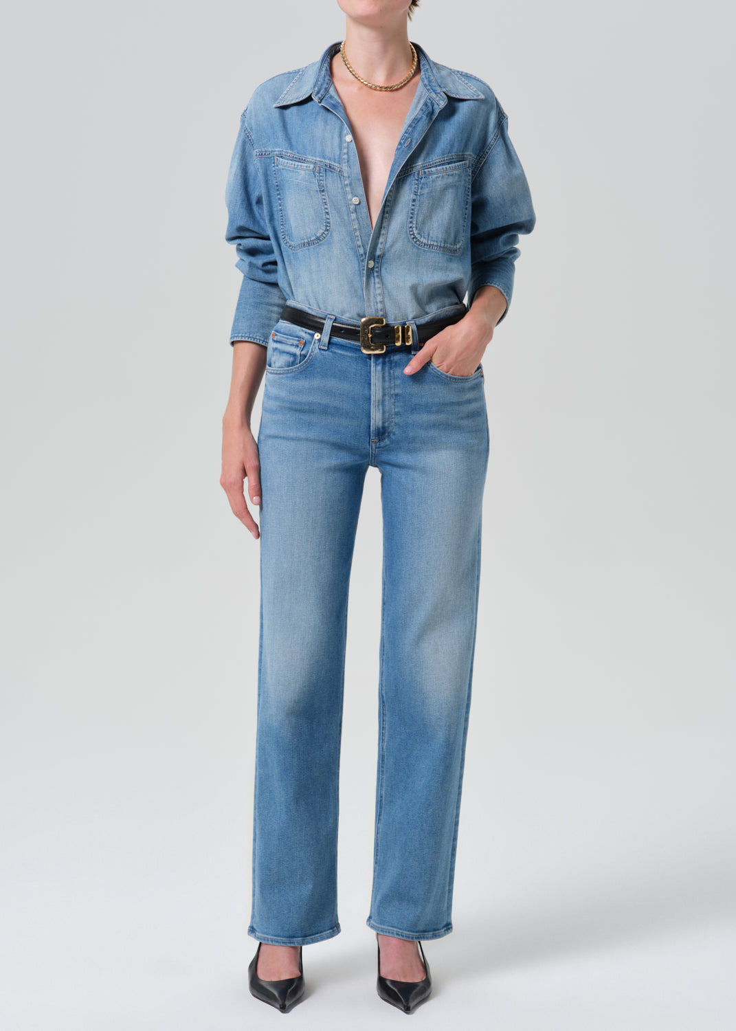 Person wearing a light blue denim shirt with light blue straight leg jeans on a grey background
