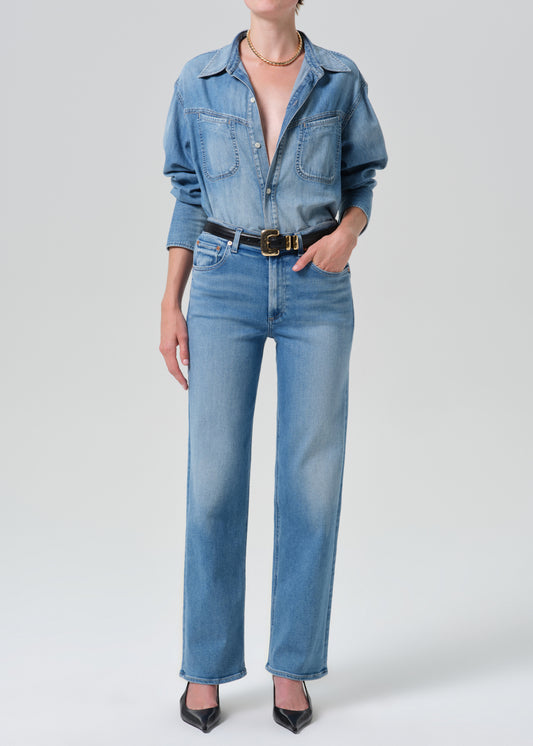 Person wearing a light blue denim shirt with light blue straight leg jeans on a grey background