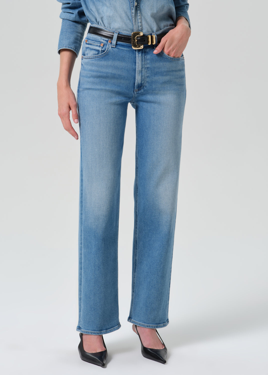 Person wearing a light blue denim shirt with light blue straight leg jeans on a grey background