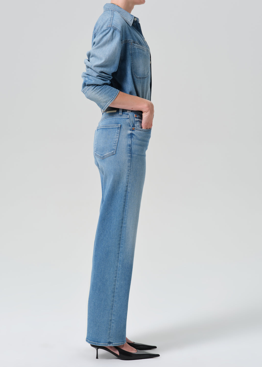 Person wearing a light blue denim shirt with light blue straight leg jeans viewed from the side on a grey background